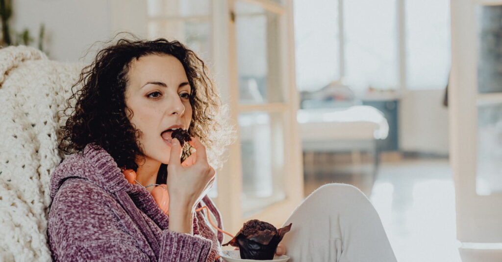Mulher comendo chocolate no sofá após o trabalho representando a vontade de doce por ansiedade e estresse.